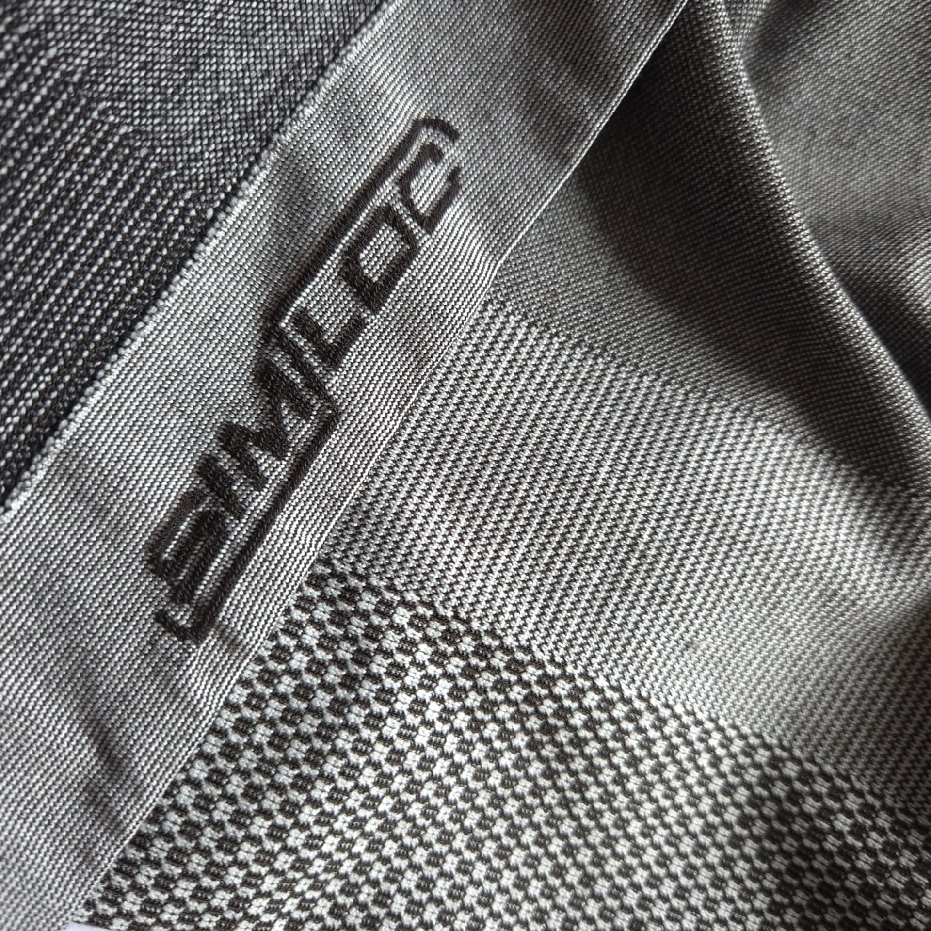 Eine Nahaufnahme des strukturierten grauen Tech-Garn-Stoffes mit geometrischen Mustern auf der ORANGE_LINE Long Shorts von HESTOR, mit "SIMLOC" in dunklem Faden an einer Kante aufgenäht.