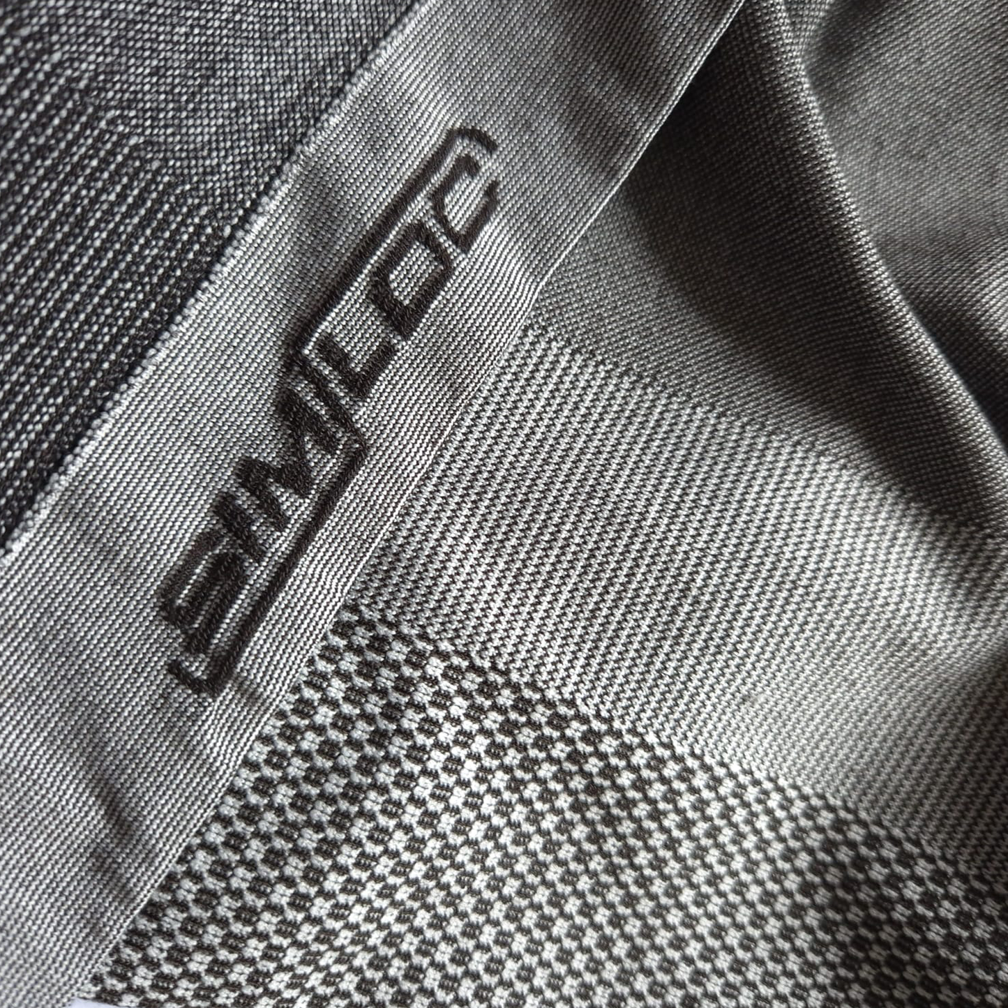 Eine Nahaufnahme der ORANGE_LINE Legging von HESTOR zeigt grau gemusterten Stoff mit "SIMLOC", der mit dunklem Garn entlang einer Kante gestickt ist, und zeigt verschiedene gewebte Strukturen und Muster.